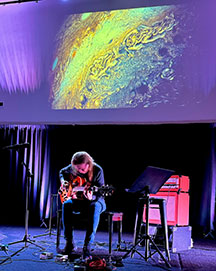 David Becker playing his guitar with a colorful slide behind him.